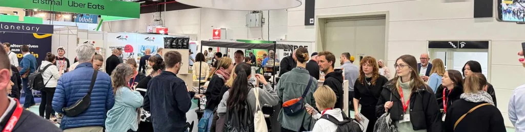Modern trade show booth in a busy exhibition hall with visible layout, back wall, lighting, graphics, visitors and circulation